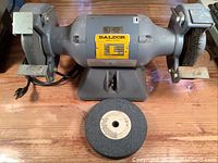 Front view of gray Baldor bench grinder with grinding wheel on left and wire brush wheel on right, placed on wooden surface with extra grinding wheel in front.