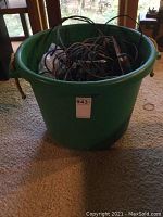 Green bucket filled with tangled multiple cables and power strips, viewed from above in a carpeted room near windows.