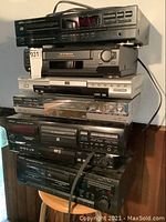 Stack of 7 electronic audio and video devices including CD players, DVD players, VHS player, cassette deck.
