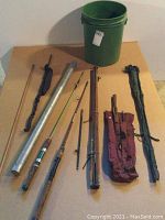 Overview of 9 fishing-related items on floor: fishing rods, rod tubes, fabric bags, green plastic bucket, wooden stick with rope loop.