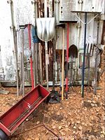 Photo showing the Toro tow behind fertilizer trailer in red metal with rust and 19 various hand yard tools standing against a metal background outdoors.