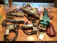 Group photo showing six power tools laid out on wooden surface including drills, sander, and grinders.