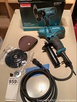 Makita GV5010 disc sander with detachable side handle, sanding disc, backing pad, power cord, and original box visible in background.