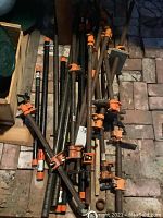Overall view of a pile of over 15 steel pipe clamps with orange handles and black pipes lying on a stone floor.
