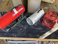 Image showing all three heaters on a table outdoors: Craftsman red gas heater, Reddy white propane heater, Marley red electric heater.