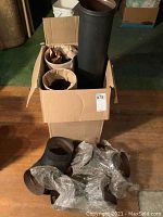 Box containing rolled metal stove pipes and three elbow joints wrapped in plastic, on wooden floor.