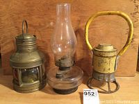 Three vintage lanterns displayed on wooden surface showing brass ships oil lantern, glass oil lamp, and yellow Adlake No. 31-F railroad lantern with labels visible.