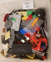 Top view of the container with assorted LEGO pieces including blocks, plates, gears, large round black pieces, and a red circular blade piece, plus an orange separator tool bagged.