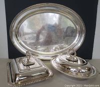 Photo showing two Birks silverplate covered vegetable dishes (oval and rectangular) placed in front of a large silverplate tray, all with decorative borders and removable lid handles on dishes.