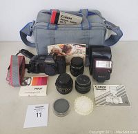 Photo showing entire lot, including Canon T70 camera, instruction manual, Speedlight 277T flash unit with case and manual, three lenses, Optex polarizer, camera bag, and small accessories arranged on table.