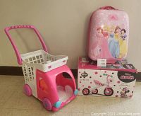 Photo of pink plastic shopping cart, Disney Princess suitcase on top of sealed Huffy Minnie Mouse pedal tricycle box