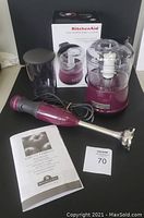 KitchenAid purple food chopper with clear BPA-free bowl, purple base, blade assembly, instruction manual, hand blender, and its power cord laid out on black surface