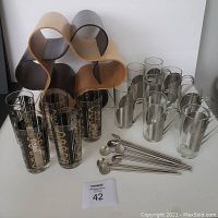 Overview: wooden wine rack, black and gold highball glasses, metal-sleeved iced coffee cups and spoons on white surface