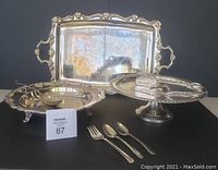 Photo showing silverplate serving tray with ornate handles, footed vegetable dish with embossed decoration, cake plate on pedestal, and assorted cutlery including spoons and fork.