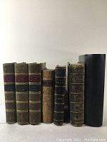 Front view of seven antique books line up showing leather and board bindings and titles on spines.