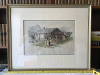 Framed watercolour painting showing a rustic farm house with mountains in the background, held in a light textured mat and simple silver frame. A measuring tape shows approximate frame size 21x25 inches.