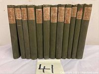 Photo of 13 hardcover books standing upright, all have green covers with faded paper labels on spines showing old edition look.