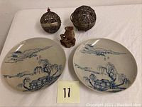 Two Canton ceramic plates with blue hand-painted landscapes, carved soapstone vase, and two ornate metal lanterns placed on a white cloth.