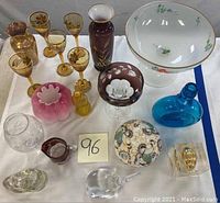 Full overview of the lot showing all colored glass items arranged on table including large rose bowl, amber cherry glasses, pink compote, powder box, blue vase, and assorted small glass items.