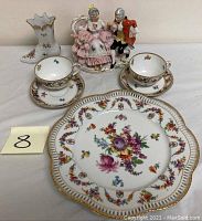 Lot of porcelain items arranged on table showing cups, saucers, reticulated plate, and figurines.