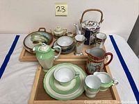Overview of porcelain lot including acorn bowl, teapot with bamboo handle, creamers, tea cups, saucers, vases arranged on wooden trays.
