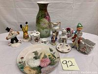 Front view of porcelain lot including vases, figurines, plate, ring holder and miniature tea cup arranged on table with lot number.