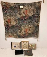Wide view of antique tapestry featuring elephants and plant motifs hanging on wall above three framed needlework art pieces. Black velvet buggy foot warmer and floral throw pillow placed on floor in front.