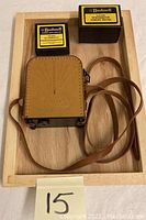 Top view of the Univex Model A camera in tan leather case with shoulder strap alongside two Bushnell camera accessory boxes on a wooden tray.