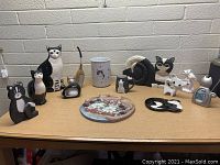 Photo shows a large assortment of cat-themed figurines and decorative items arranged on a table in front of a brick wall. Different materials include wood, ceramic, and metal. Various styles from realistic to abstract.