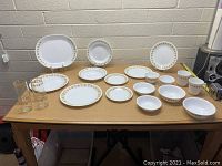 Large set of vintage Corelle dishes including plates, bowls, and glasses arranged on a wood table against a beige wall.