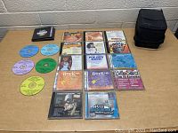 Photo showing all 19 karaoke CDs spread out on a table alongside a black soft-sided carrying case.