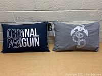 Photo showing lot of two new throw pillows, one navy blue with text and one grey with penguin graphic.