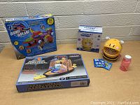 Lot of 3 kids toys on a wooden surface against a cinder block wall, showing the Bionic Biplane box, Colorful Bubble Bee bubble blower, and Head to Head Basketball game box.