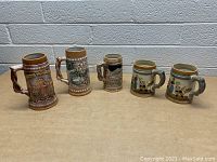 Front view showing five distinct decorative steins and mugs on a surface against a brick background.