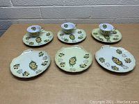 Six Lefton china cups, saucers, and oval plates displayed on wooden surface against brick wall. All items feature yellow flower design with green leaves.