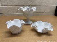Three vintage white milk glass hobnail pattern pieces displayed on table against beige brick wall: pedestal bowl, small vase, shallow dish with ruffled edges