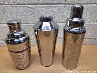 Photo showing three stainless steel cocktail shakers with varied designs placed side by side on a wooden surface against a rough beige wall.