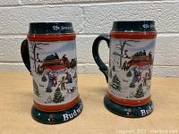 Pair of 1991 Budweiser collectible steins featuring winter holiday scenes with horse-drawn Budweiser wagon, people, and snowy landscape. Green base and top rim with 'Budweiser' and 'The Season's Best' text.
