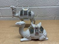 Pair of white ceramic camels with detailed patterned saddles, one standing two-hump camel and one lying one-hump camel.