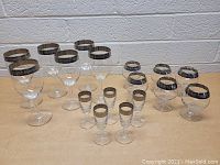 Full lot of 15 glassware pieces arranged with front row containing small cordial glasses, middle row with lowball glasses, and back row with taller wine glasses. Each glass has a dark decorative rim with intricate patterns.