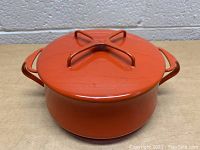 Front view of vintage Dansk Kobenstyle orange enamel stock pot with lid on wooden surface against gray brick wall.