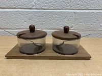 Front view of the pair of condiment servers with wooden lids and spoons on a wooden tray.