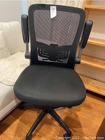 Front view of black mesh back office chair with folding padded arms, cushioned seat, and rolling casters on hardwood floor.