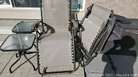 Two beige folding loungers next to two small square tables with glass tops outside on patio.