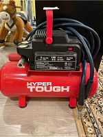 Front view of the red Hyper Tough compressor with brand and tank size visible, and blue hose wrapped around handle.