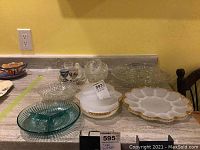 Several glass plates, bowls and egg plates displayed on counter including two white milk glass egg plates with gold trim, clear glass bowls and decorative glass plates.