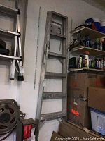 Aluminum folding ladder upright against wall with several cardboard boxes and shelves nearby.