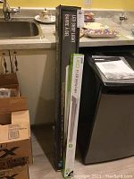 Photo showing two boxed LED lights standing upright next to a kitchen counter, one taller and wider box labeled 'LED Shop Light Barrea Del' and the other smaller and thinner labeled '3 ft LED Utility Light'.