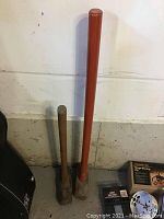 Two sledgehammers standing upright against a wall, one with a long orange handle and another smaller wooden handled sledgehammer.