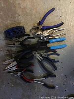 Overview of the assorted pliers laid out on a concrete floor with some pliers stacked overlapping.